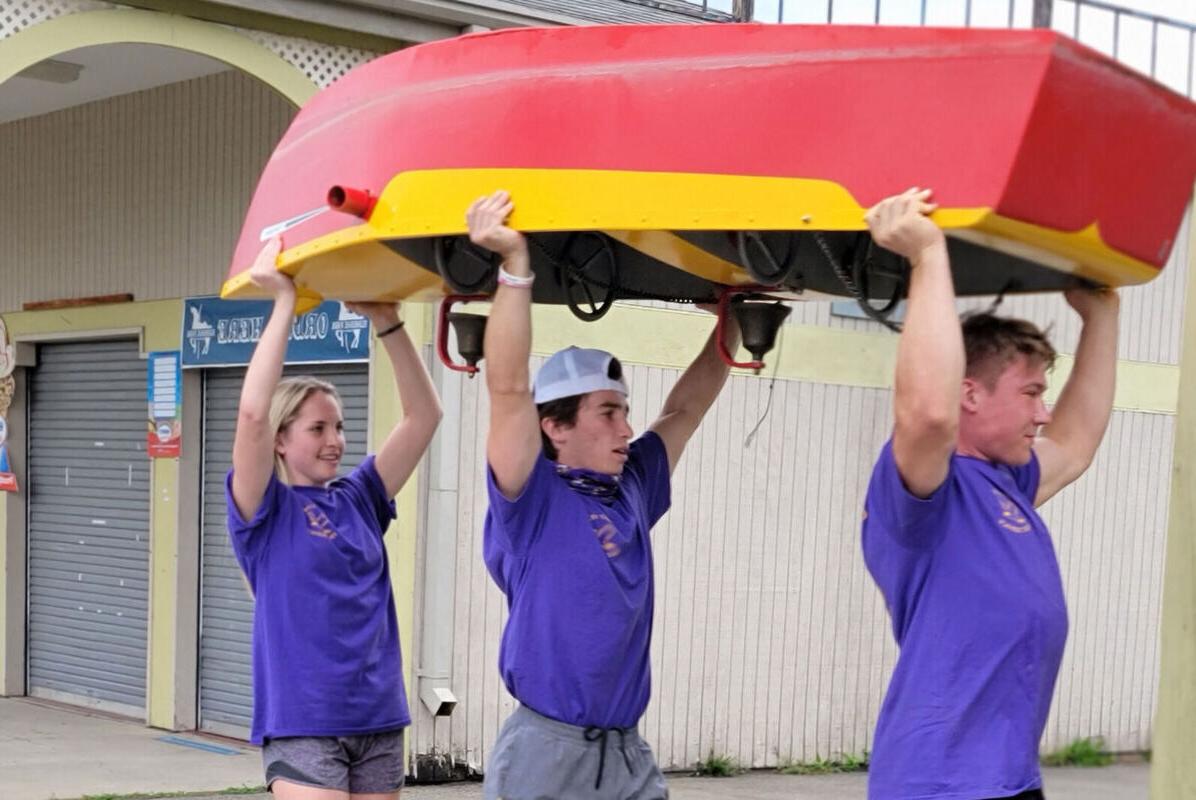 Two male students and a female student carry a boat above their heads across Eldridge Park during a community service outing